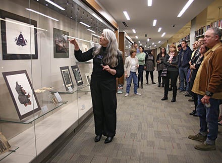A curator from the University Art Collection points at an artwork on exhibit during a gallery event as a crowd of onlookers gather around her
