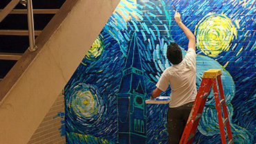 Agree Ahmed, a male student, is shown from behind perched atop a ladder as he adds brushstrokes to a mural depicting Healy Hall in the style of Van Gogh's "A Starry Night" with swirling blues and yellows symbolizing the night sky, stars, and streetlights.