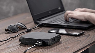 A small black power bank sits next to a laptop, phone, and sunglasses as the hands of an unseen person types on the laptop, which is being charged by the power bank.