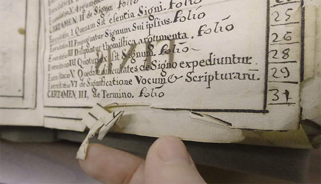 book page with handwritten Latin text with paper breaking at corroded ink border and held by a person's two fingers before conservation treatment