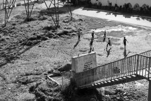 Black and white photo of several children playing soccer at the base of long concrete steps in an empty lot of land. In the background is the lower portion of a large tower.