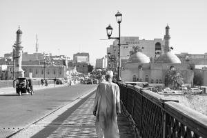 The photo shows man in traditional Iraqi clothing walking away from the camera across a bridge. 