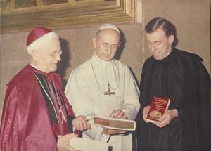 Three clergy, the cardinal in red robes on the left, the pope in white robes in the middle, and a priest on the right in black. The pope holds a book in his hands. 