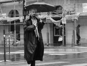 Black and white photograph of a man with an umbrella who is wearing a black raincoat walking across a city street
