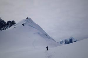 Color photograph of a snowy, mountainous landscape, with a woman in the center and footsteps leading up to her. Taken in Antarctica on the Melchior Islands. 