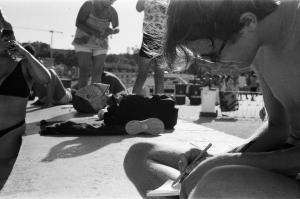Black and white photograph of a few human figures seen in blur. The only person in focus is a boy on the right side, seen from the side. He is shirtless, sitting and hunched, holding a pencil to paper. There appears to be a city skyline in the background. 