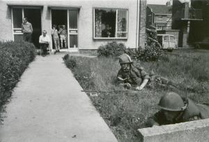 Photograph of a solider laying prone on a lawn with onlookers in the background