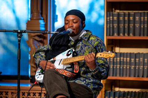 performer wearing a black head scarf and a decorative blue and green shirt playing a guitar and singing into a microphone