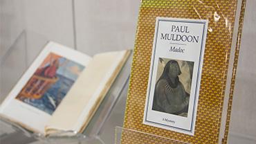 A copy of Paul Muldoon's "Madoc" displayed next to another open book in an exhibition shelf.