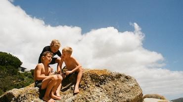 Color photography of three boys sitting on a large rock. There are clouds and a blue sky in the background.