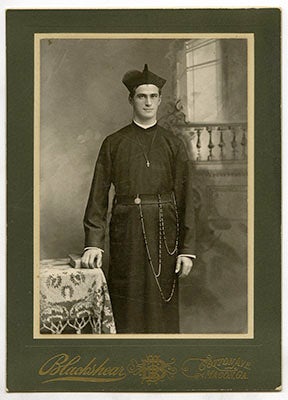 Daniel Cronin, S.J. stands in his cossack, his right hand resting on a book on a table next to him, the name of the Blackshear photo studio printed in gold below the photo