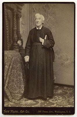 James Curley, S.J., standing in his cossack while leaning on a cane in his right hand and his left hand tucked in a hand-in-waistcoat pose, with the name of the photography studio New Photo Art Company printed below the photo