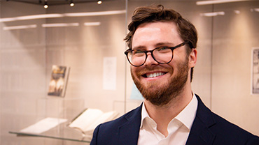 Chris Kenny, a white man in his 20s with brown hair, glasses, and beard, smiles in front of an exhibition with open books and art.