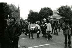 Black and white photograph of protesters outside the main gates, some of whom are pushing a white sedan towards the gates