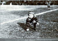 A black and white photograph of a small boy dressed in a football uniform taking a three point stance on the field.