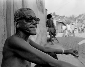 Black and white photo of a man sitting on the sidewalk wearing dark sunglasses. He is in semi profile and shown from the torso up shirtless.