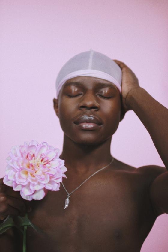 A shirtless black man wearing a white skull cap and a necklace poses with his eyes closedand his left hand touching his head while his right hand holds a large pink flower