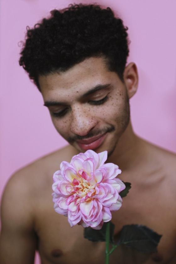 A shirtless black man with beard and moustache bows his head towards a large pink flower with his eyes closed