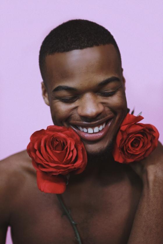 A shirtless black man laughs with his eyes closed and two red roses wrapped around his neck