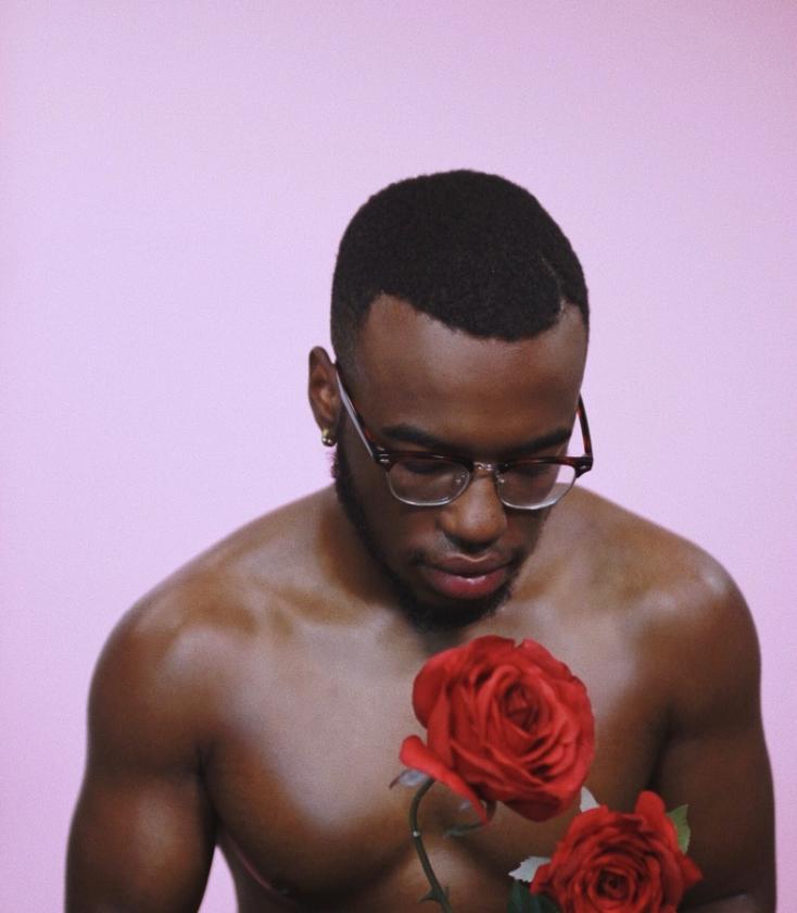 A shirtless black man wearing glasses looks down at two red roses with his eyes closed