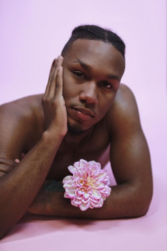 A shirtless black man looks at the camera while resting on his left elbow and with his right hand cupping the right side of his face, while a large pink flower rests on his left forearm