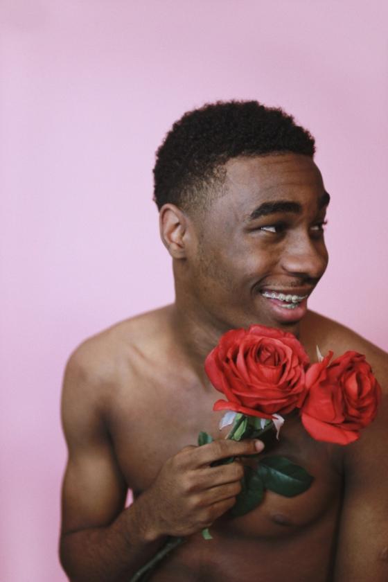 A shirtless black man looks to his left and smiles while holding two red roses in his right hand