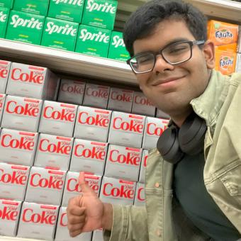 Arun Lakshman smiles and gives a thumbs up hand gesture in the sode aisle of a grocery store in front of a Diet Coke display