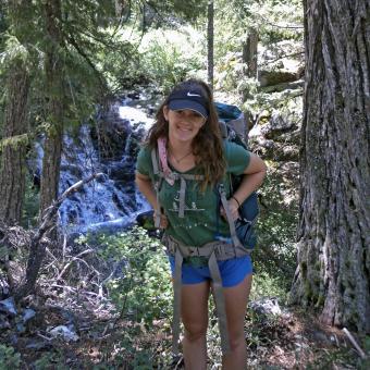 Emily Kaney smiles while carrying a large backpack in front of a waterfall in a forest