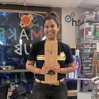 Nikisha Kotwal stands in the Maker Hub wearing a Garfield t-shirt and holding a wooden clock with a gingko leaf motif