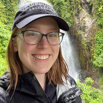 Olivia Geist wears a rain jacket and a Georgetown ball cap while standing in front of a waterfall