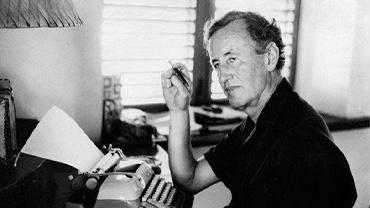 Ian Fleming sitting at his typewriter, looking off into the distance.
