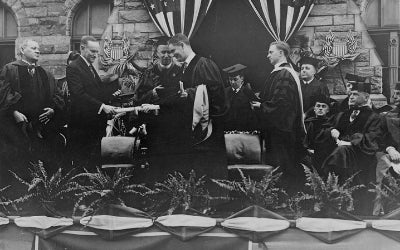 President Calvin Coolidge presenting an honorary degree to Governor William S. Flynn of Rhode Island at the Georgetown University commencement ceremony. To the left of President Coolidge stands Pierce Butler, Associate Justice of the Supreme Court. To his right, stands Georgetown President John B. Creedon, S.J.